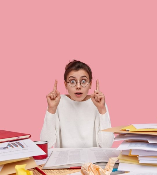 Photo of surprised young woman works at desktop, shows direction upwards, indicates with fore fingers, amazed to hear noise upstairs, reads textbook, works with documents, isolated on pink wall